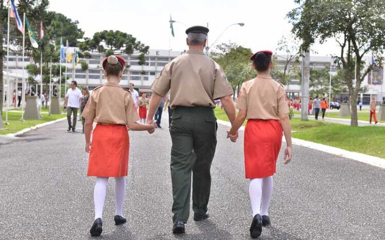 Um militar levando pelas mãos duas alunas do Colégio Militar em foto de costas.